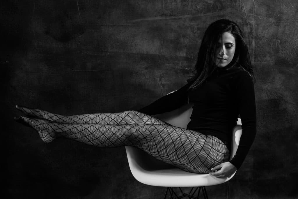 Woman with dark long hair sitting on a chair with her legs in the air, wearing a black turtleneck and fishnet stockings