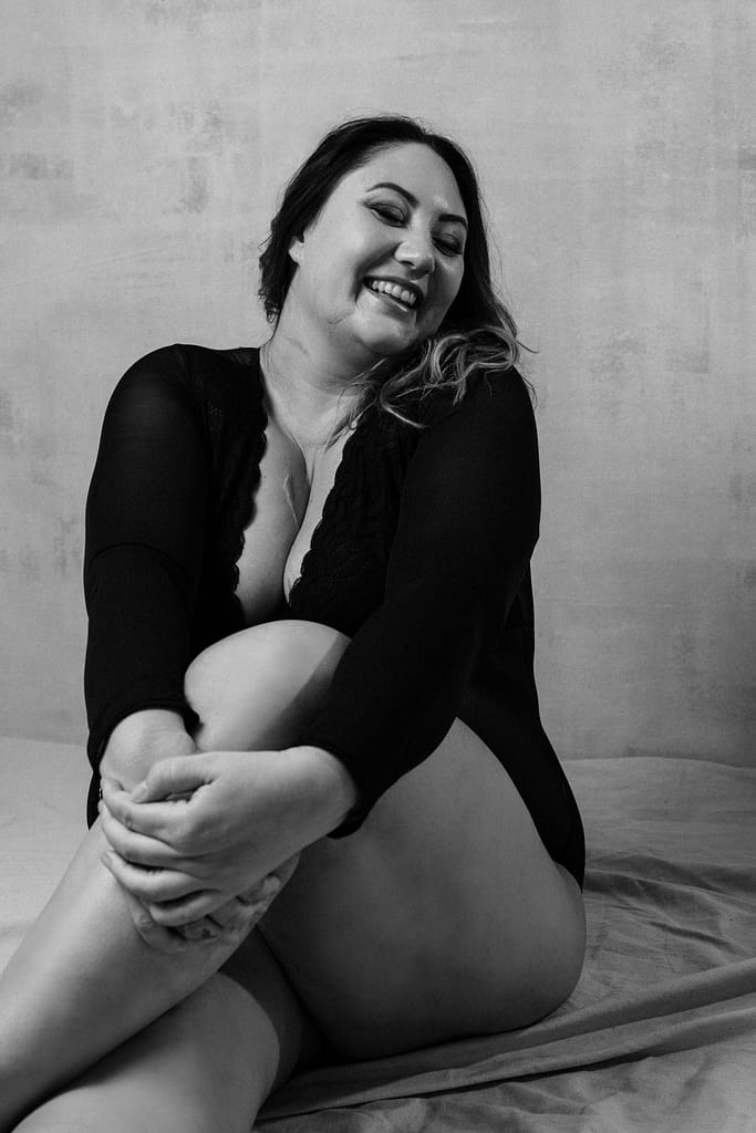 Vancouver Plus Size Boudoir, showing a woman in a black long sleeved bodysuit with a big smile on her face in a contemporary boudoir studio.