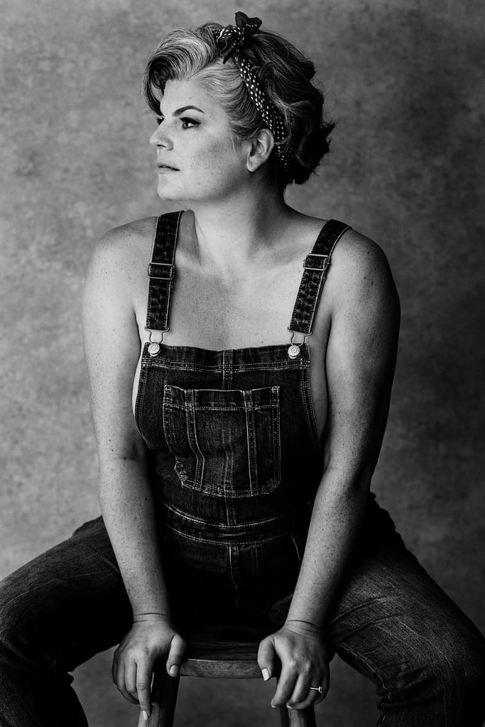 black and white boudoir photoshoot image of woman sitting on stool wearing only overalls