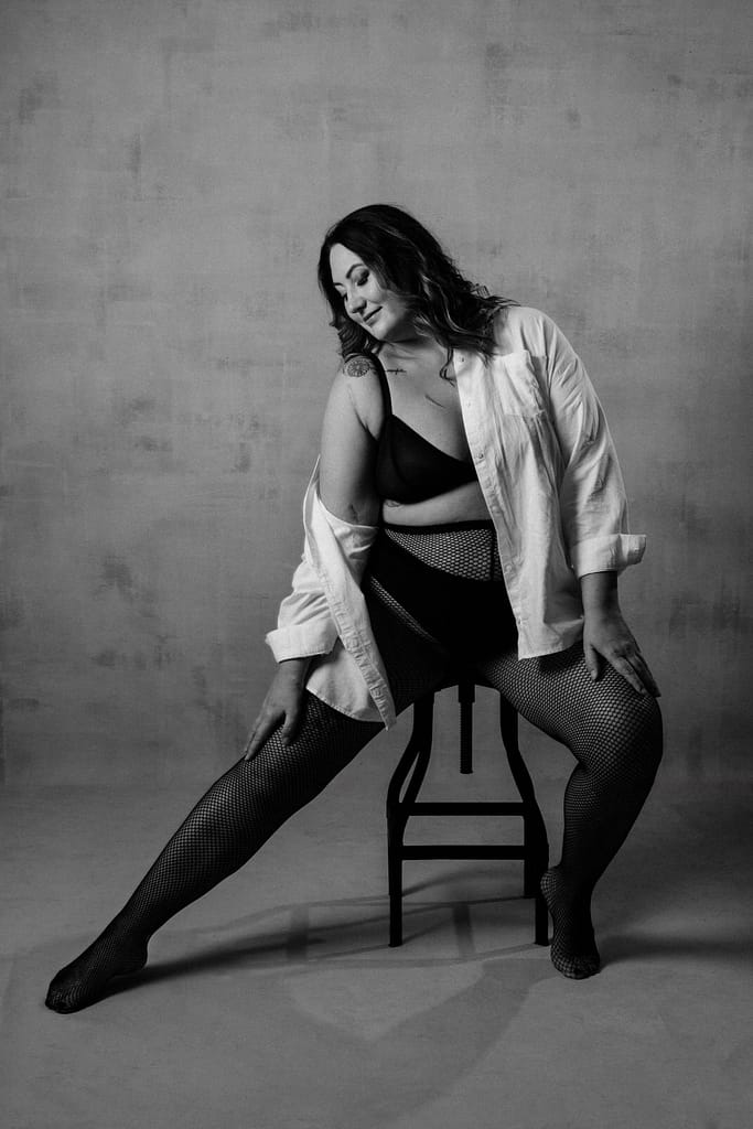 Vancouver Plus Size Boudoir, showing a woman in a black fishnets and a white blouse with a black bra offering a classic timeless look with a smile on her face in a contemporary boudoir studio.