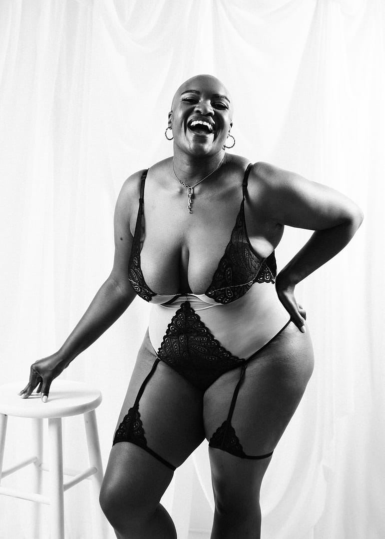 vancouver boudoir photographer image of plus size black woman with alopecia wearing lingerie and leaning on stool while laughing