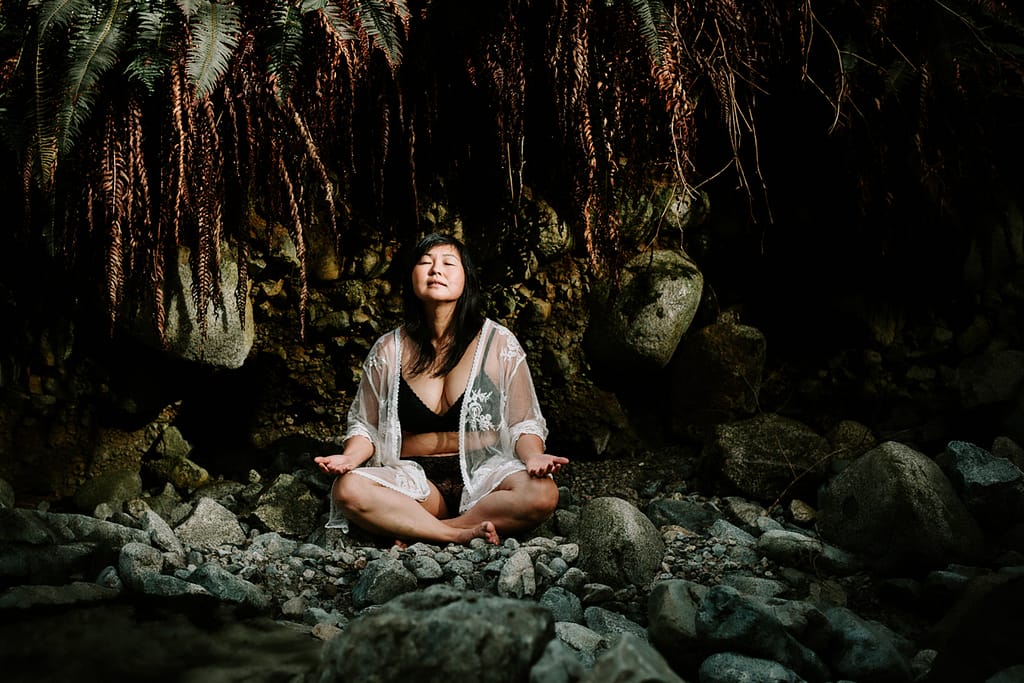 woman sitting crossed legged with eyes closed and open palms to the sky laid on her knees outdoors in a forest 