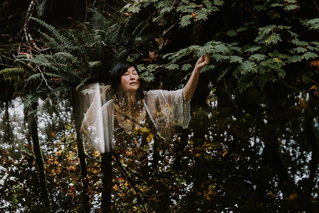 creative outdoor boudoir image in colour with green leaves, tree trunks and sky peeking through with woman in the centre of image in a white lace shawl