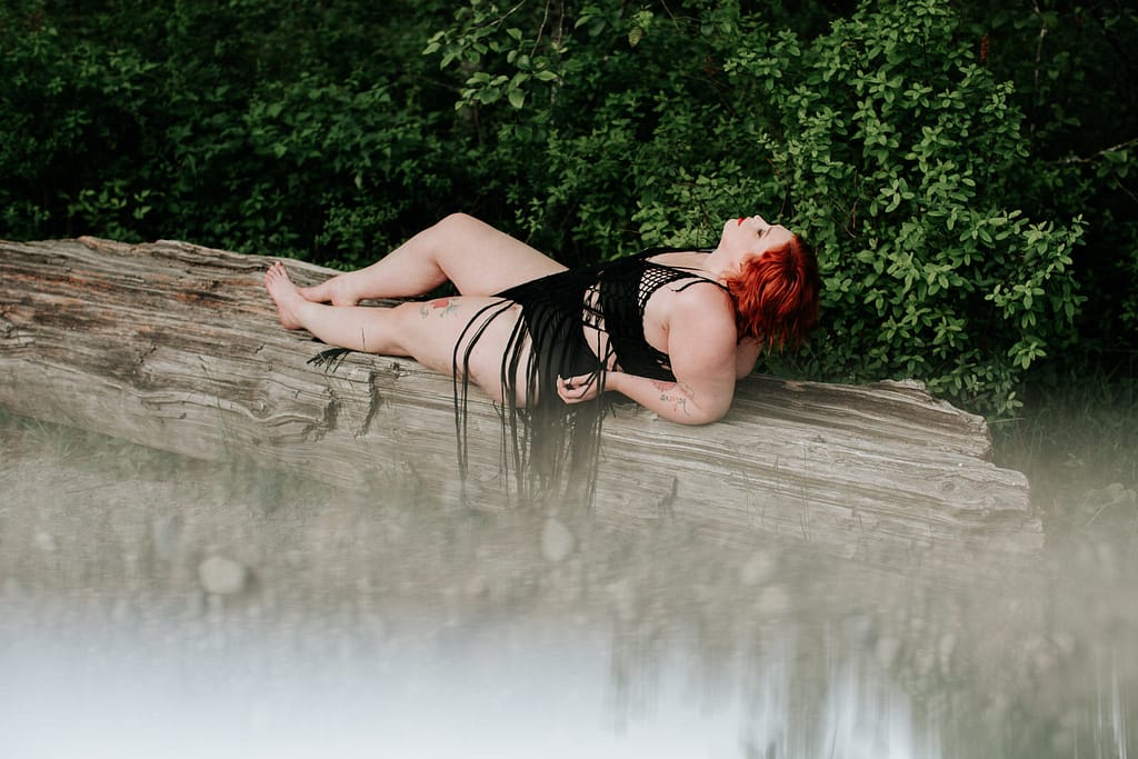 woman with red hair and wearing a black with trim bikini laying on her back on a log with bushes behind her