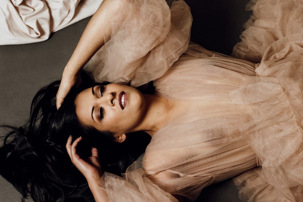 Woman laying on her back with her hands at her head entwined in her black hair and wearing a blush coloured sheer gown