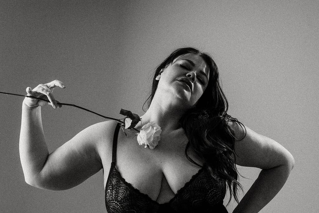 Plus size boudoir photo of a woman tilting head holding a rose to her decoletage