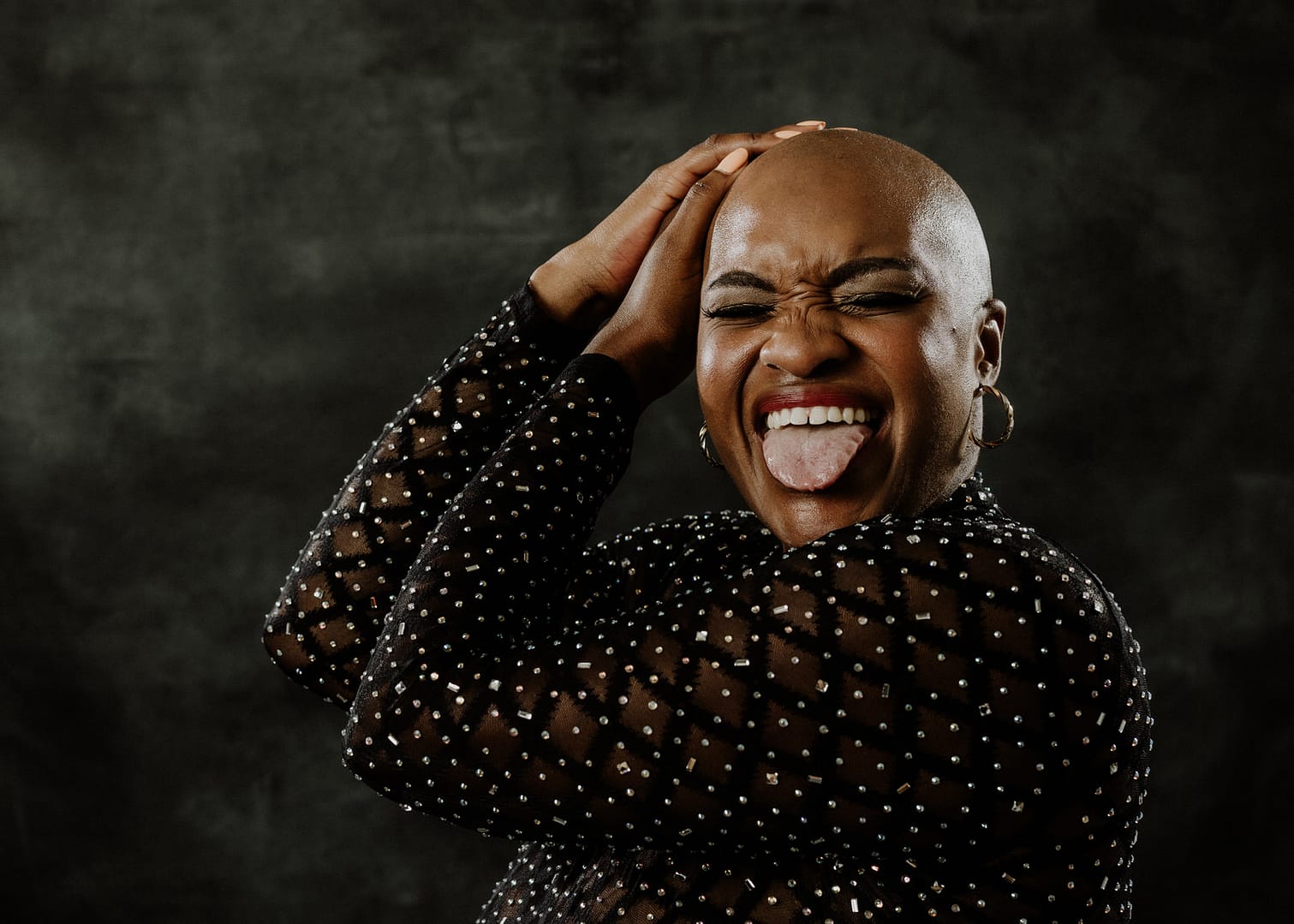 boudoir photo of plus size black woman with alopecia sticking out tongue hands on head