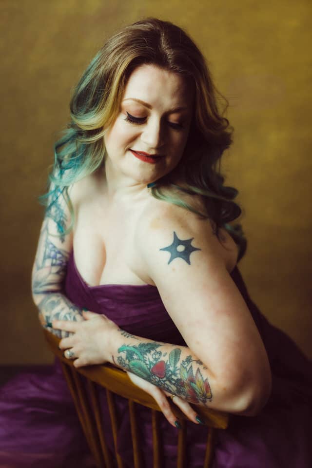 A tattooed woman with rainbow hair wearing a strapless gown sitting backwards on a chair