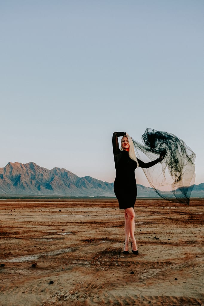 Blonde haired woman wearing a black dress and heels holding black fabric standing in the Las Vegas desert with mountains in the background