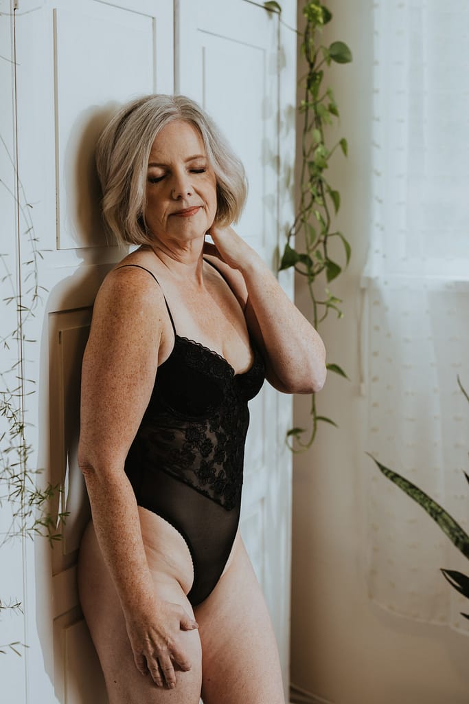 A woman wearing a revealing black lace body suit leaning against a boudoir with her hand up at her neck. Bonnie is 40 over 40