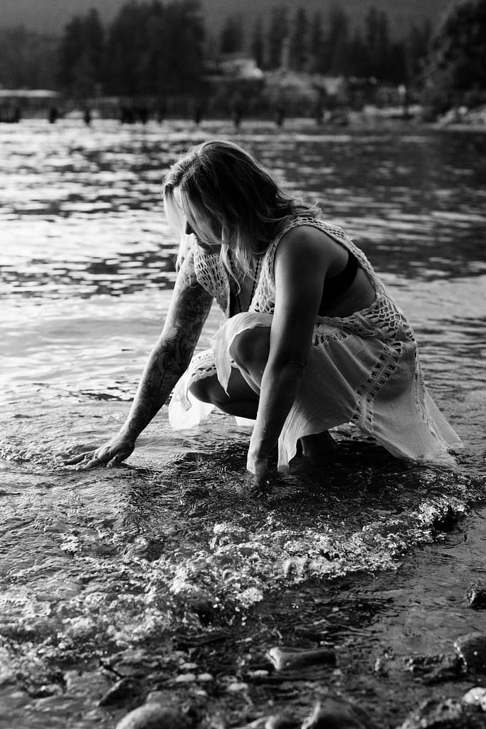 a fabulous woman over 40 with her feet in the ocean, she's kneeling down with her hands and bottom of her dress in the water