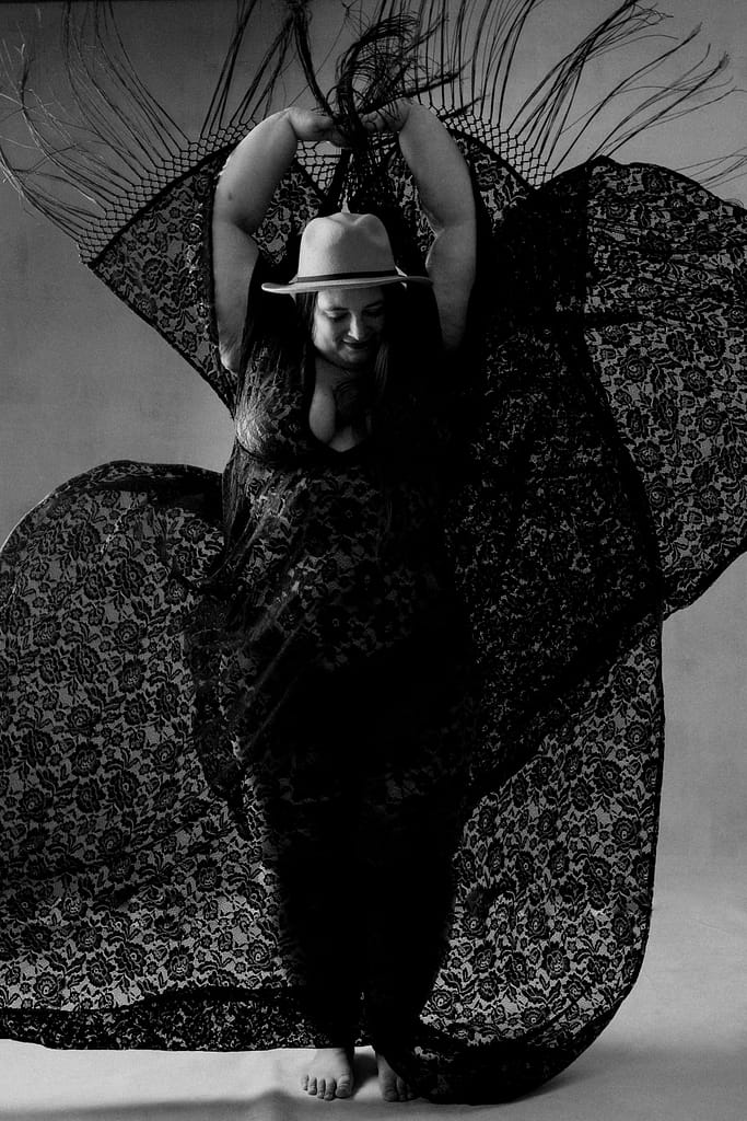 black and white boudoir image of woman wearing lacey robe and hat and arms in the air