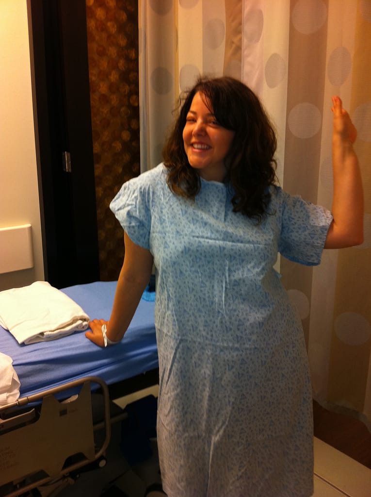 Female patient wearing a hospital gown in a hospital room standing beside a hospital bed waving before getting an angio