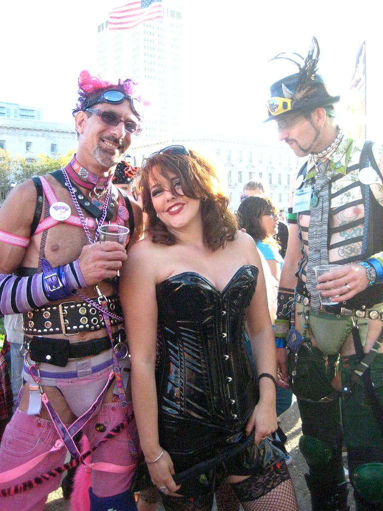 Woman in sexy PVC bustier and fish nets flanked by two men in alternative Love Parade attire