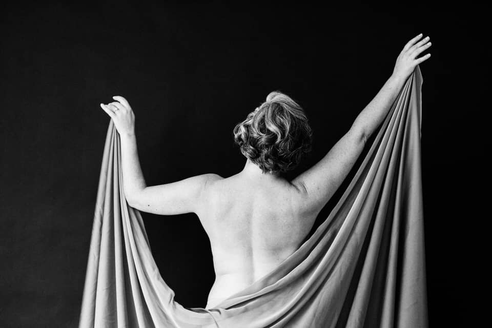 a woman standing with back facing camera and holding up fabric revealing her bare back and arms in boudoir photoshoot
