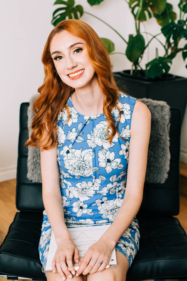Woman with red hair sitting on a black chair wearing a blue and white floral dress
